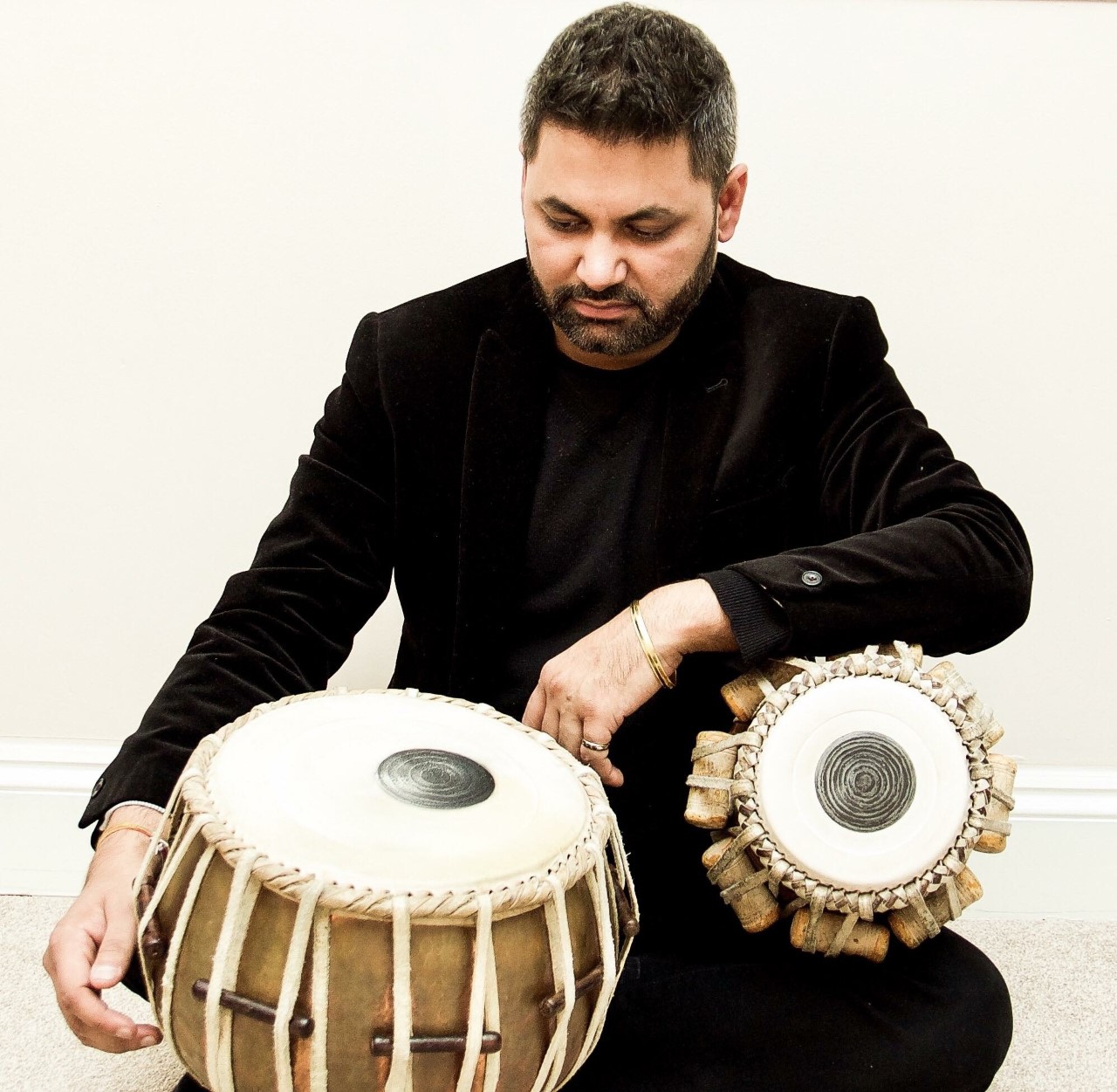Alok Verma, seated and wearing all black, with two wooden tabla drums
