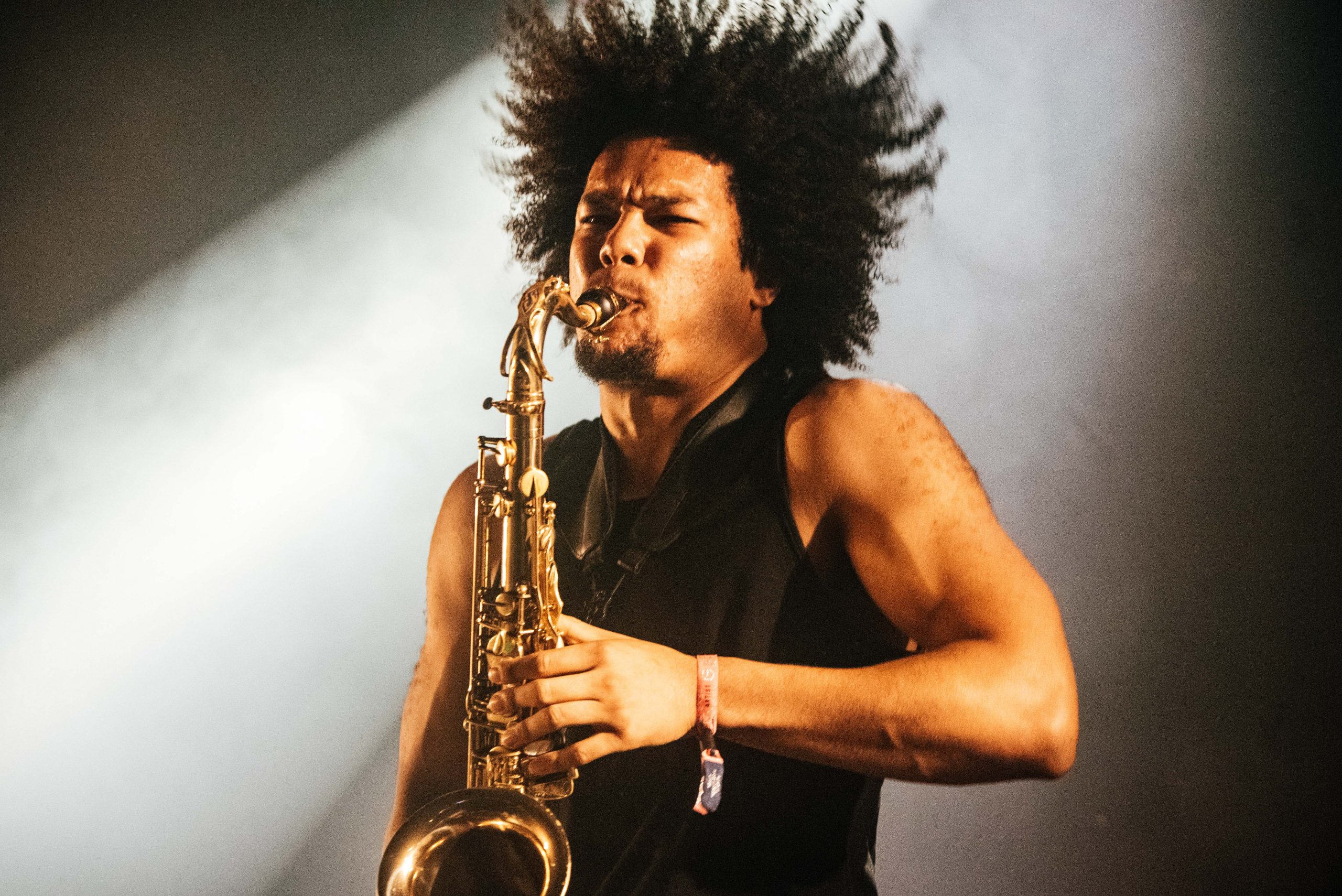 Kaidi Akinnibi, in a black vest top playing his saxophone on stage with gusto, a white spotlight shining down behind him
