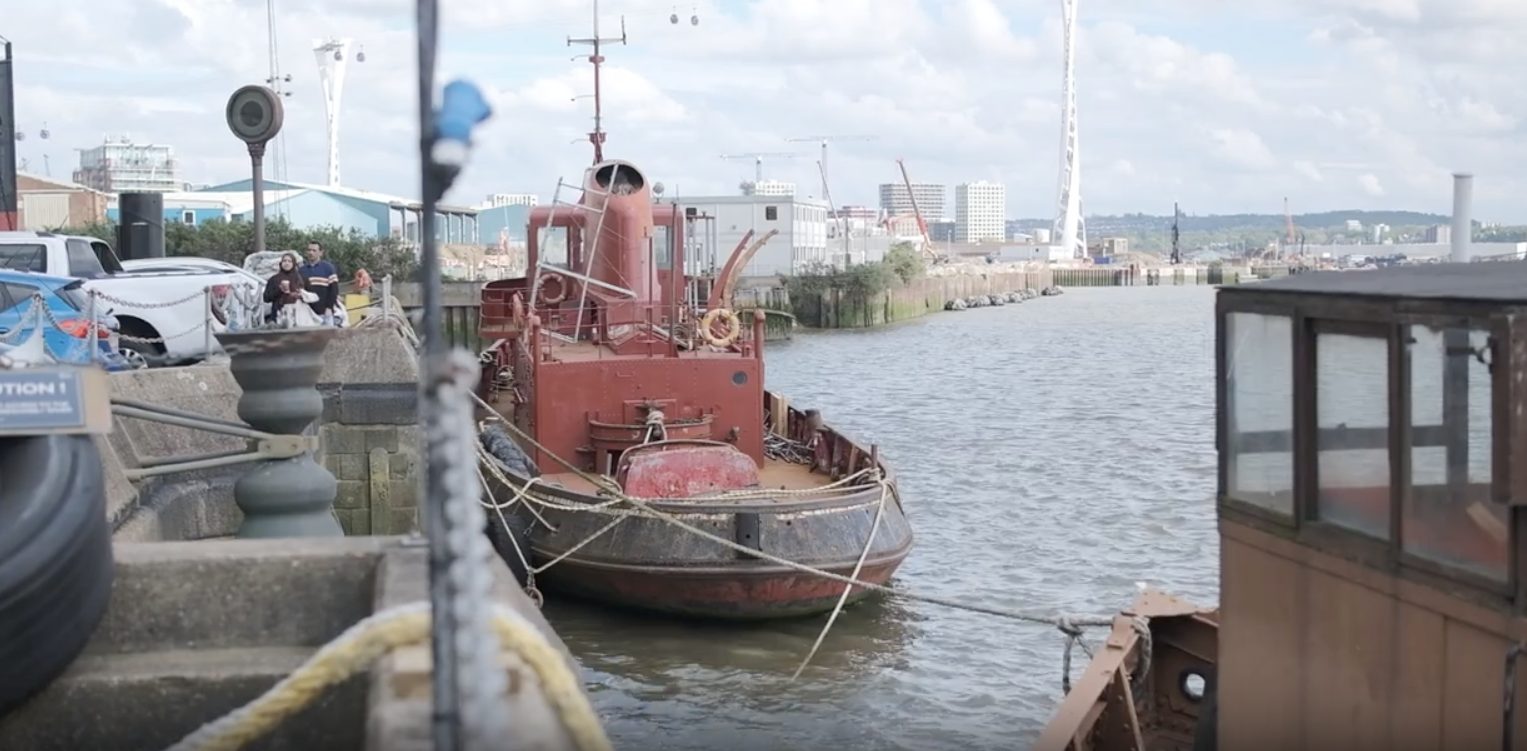 Image shows boat at Trinity Buoy Wharf with buildings and people walking along