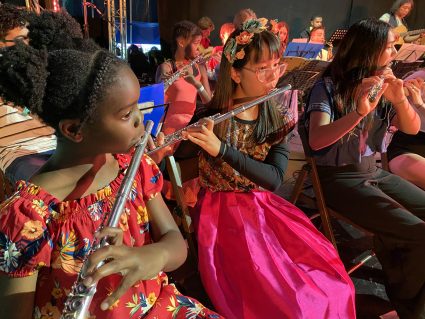 A group of colourfully dressed young musicians, seated playing flutes.